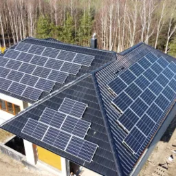 Widok z góry na dom z panelami słonecznymi na dachu, logo firmy w lewym górnym rogu, teren wokół domu częściowo przygotowany pod zagospodarowanie.