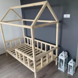 Surowa drewniana rama łóżka w kształcie domku, z listwami pod materac, ustawiona w pokoju z szarą ścianą i dwoma białymi lampionami dekoracyjnymi obok.