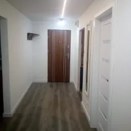 Nowoczesny przedpokój z panelami podłogowymi w odcieniach szarości, białymi ścianami i drzwiami wewnętrznymi z przeszkleniami, oświetlony listwą LED w suficie.