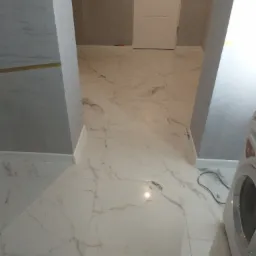 Białe marmurowe płytki podłogowe z szarymi żyłkami, szara ściana, białe drzwi w tle i fragment pralki po prawej stronie.