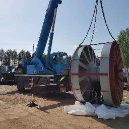 Niebieski dźwig podnosi element konstrukcyjny wentylatora przemysłowego na zewnątrz, w tle widoczny las i budynek.