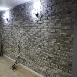 Ściana wykończona dekoracyjną okładziną imitującą cegłę, oświetlona dwoma kinkietami, z widocznymi kablami na podłodze i fragmentem mebla w tle.