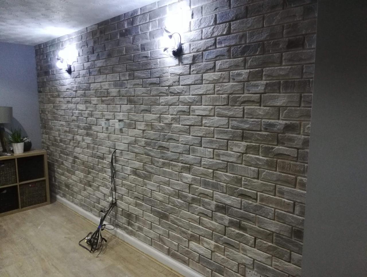 Ściana wykończona dekoracyjną okładziną imitującą cegłę, oświetlona dwoma kinkietami, z widocznymi kablami na podłodze i fragmentem mebla w tle.