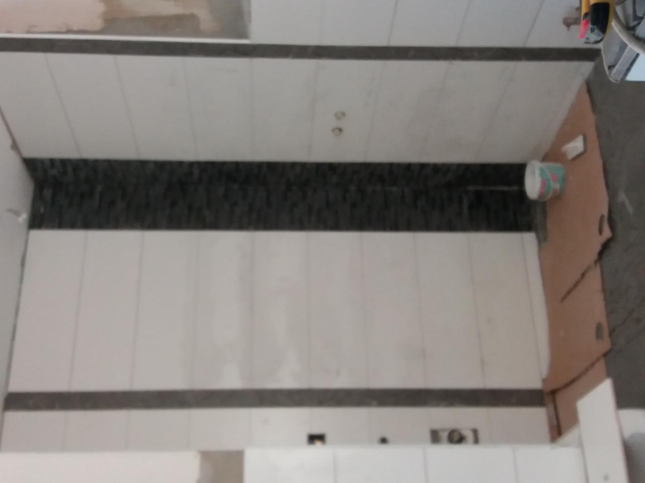 Ściana w trakcie układania płytek, widoczny pas dekoracyjny z ciemnych płytek, przygotowane miejsce na gniazdka elektryczne, zabezpieczona folią część ściany.