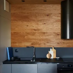 Nowoczesna kuchnia z drewnianą ścianą nad blatem, czarnym okapem i szarymi szafkami. Na blacie deski do krojenia i minimalistyczna armatura.