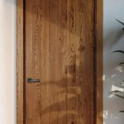 Drewniane drzwi wewnętrzne z pionowym usłojeniem,  ciemna klamka, minimalistyczna ościeżnica, w tle roślina doniczkowa i ściana w neutralnym odcieniu.