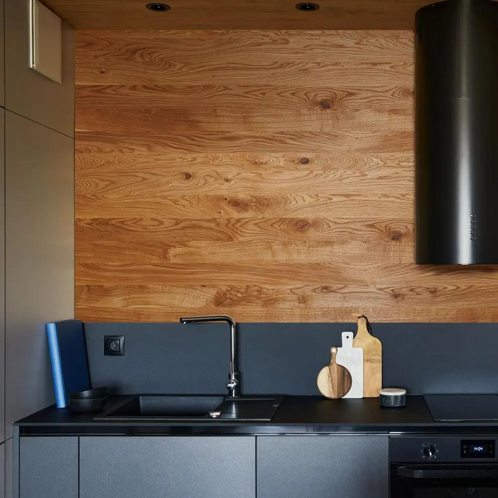 Nowoczesna kuchnia z drewnianą ścianą nad blatem, czarnym okapem i szarymi szafkami. Na blacie deski do krojenia i minimalistyczna armatura.