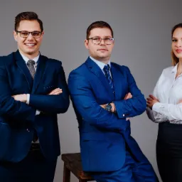 Trzy osoby w garniturach i koszuli na szarym tle, dwoje z nich ma założone okulary, jeden mężczyzna siedzi na drewnianym stołku.