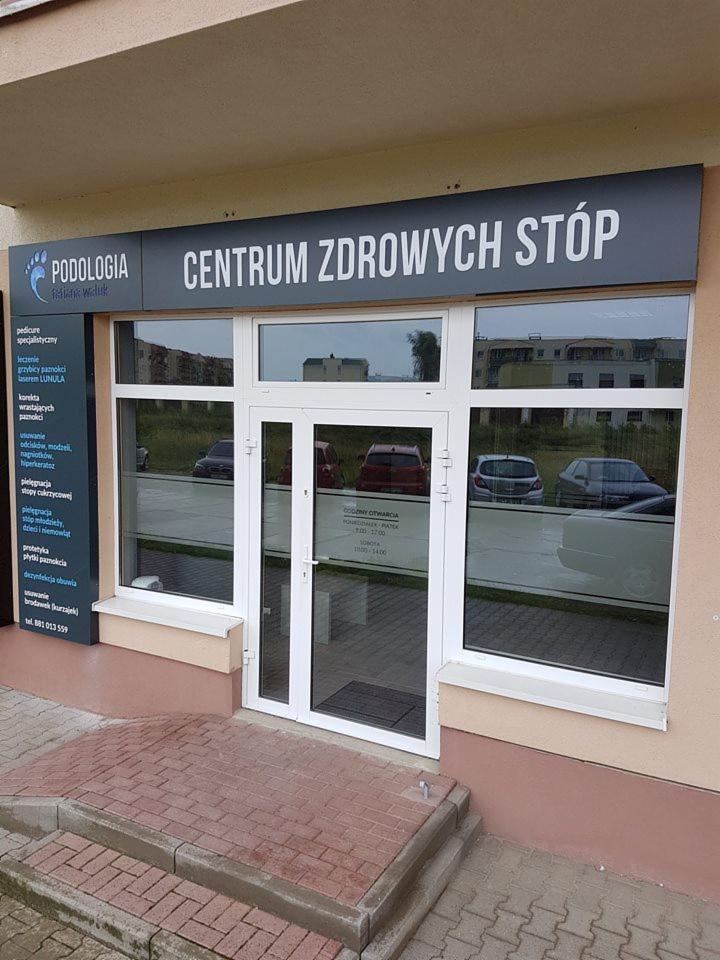 Fasada budynku z szyldem 'Podologia - Centrum Zdrowych Stóp', widoczne wejście z białymi drzwiami i okna z odbiciem samochodów.
