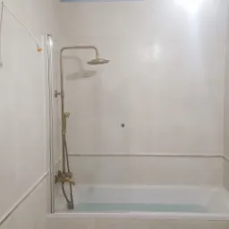 Jasna łazienka z wanną, kabiną prysznicową z mosiężną armaturą i grzejnikiem drabinkowym; widoczne niedokończone podłączenie elektryczne nad kabiną.