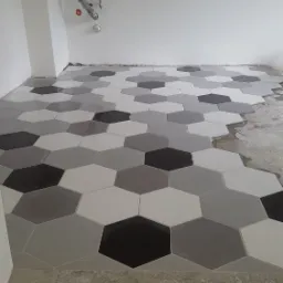 Fragment podłogi łazienki z ułożoną, heksagonalną mozaiką w odcieniach szarości i bieli, widoczne rury instalacji wodnej na ścianie oraz fragment surowej posadzki w tle.