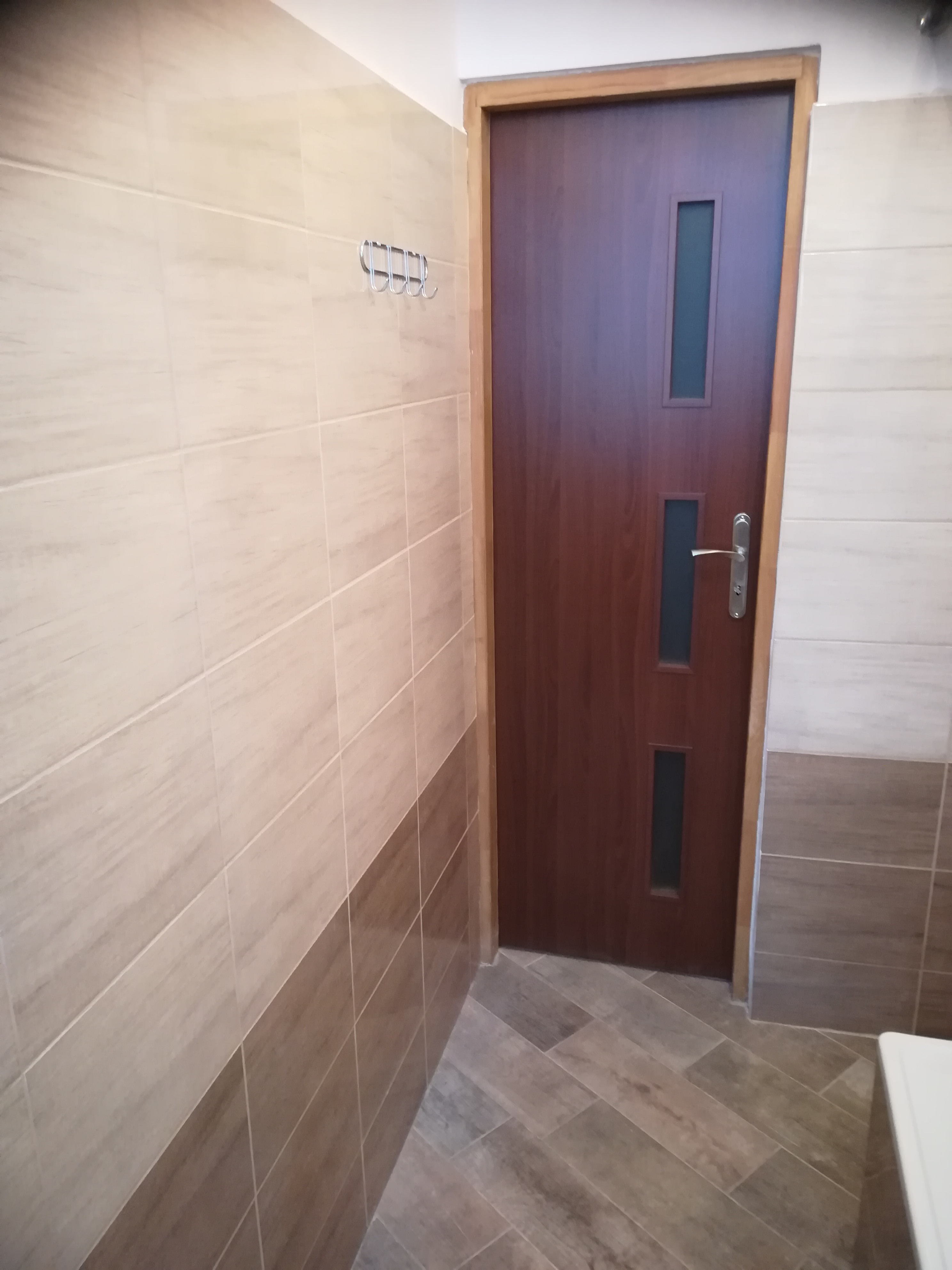 Wykończona łazienka z beżowymi płytkami ściennymi i podłogowymi imitującymi drewno, brązowe drzwi z szybkami i chromowane wieszaki na ścianie.