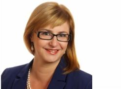 Portret uśmiechniętej kobiety w okularach, blond włosach i niebieskiej marynarce na białym tle.