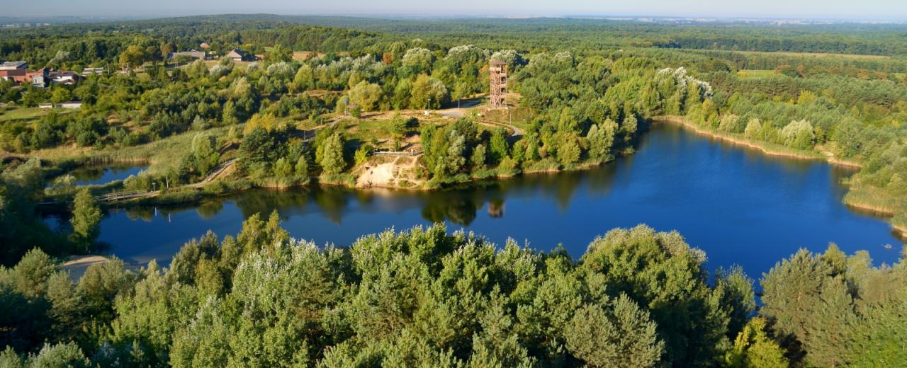 Panoramiczny widok z lotu ptaka na jezioro otoczone lasem, z drewnianą wieżą widokową na wzgórzu i budynkami w oddali.