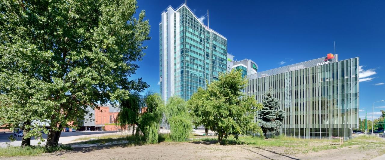 Nowoczesny biurowiec z zielonymi elementami architektonicznymi, widoczny spod drzew z liśćmi, w słoneczny dzień.