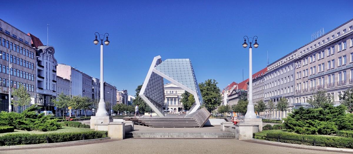 Nowoczesna szklana instalacja artystyczna na placu miejskim w Poznaniu, otoczona klasycystycznymi budynkami i latarniami.