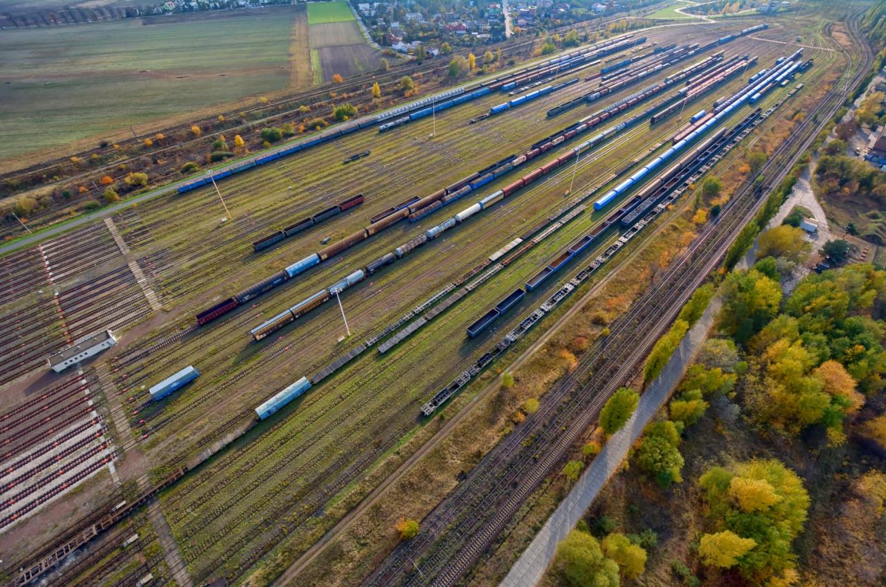 Szeroki plan z lotu ptaka na stację kolejową z wieloma torami i wagonami towarowymi, zalesiony obszar z jesiennymi kolorami drzew po prawej stronie, oraz pole i zabudowania w tle.
