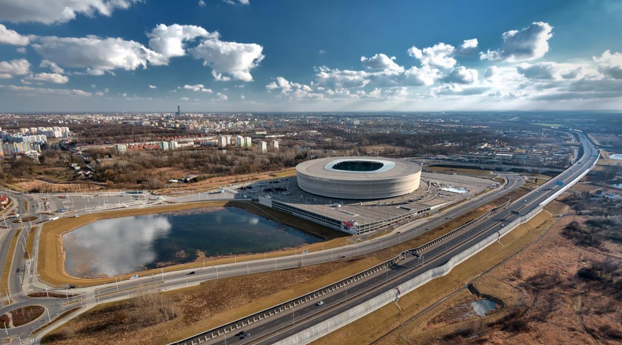 Panorama miasta z perspektywy lotniczej, z widocznym stadionem w kształcie pierścienia, jeziorem odbijającym chmury i rozległą infrastrukturą drogową.