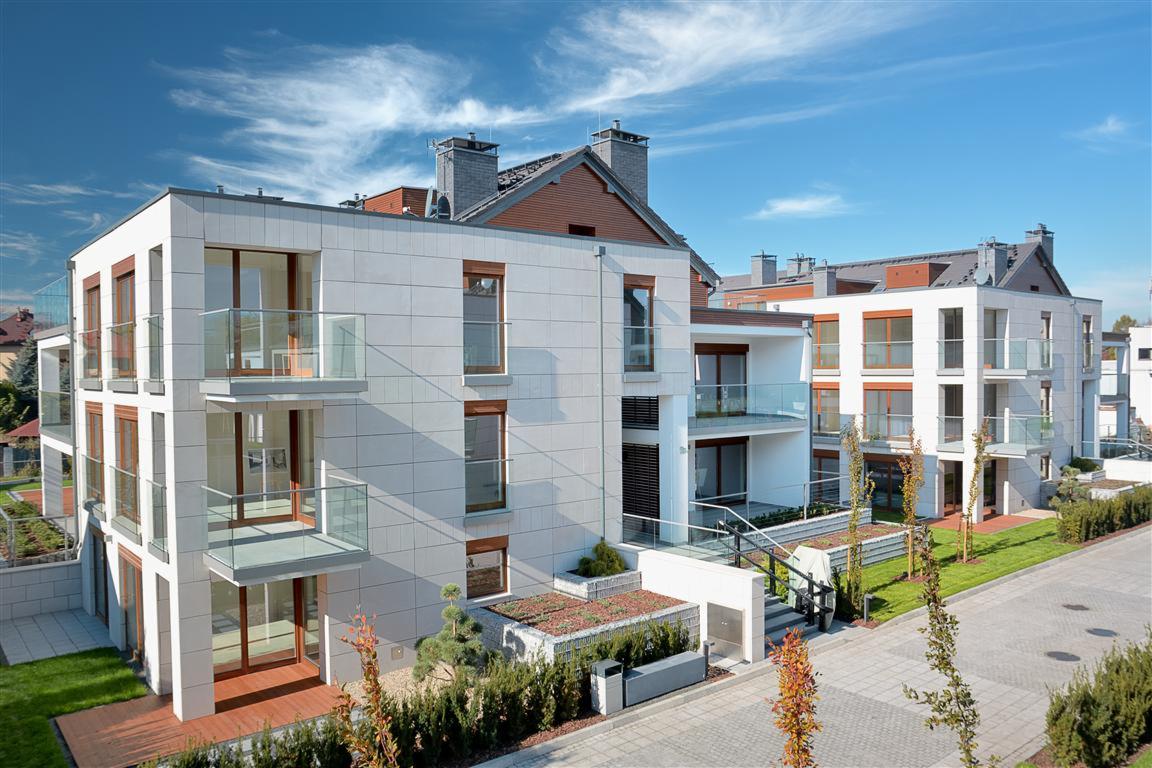 Nowoczesny budynek apartamentowy z białymi elewacjami, szklanymi balkonami i zadbanym otoczeniem, widok z zewnątrz w słoneczny dzień.