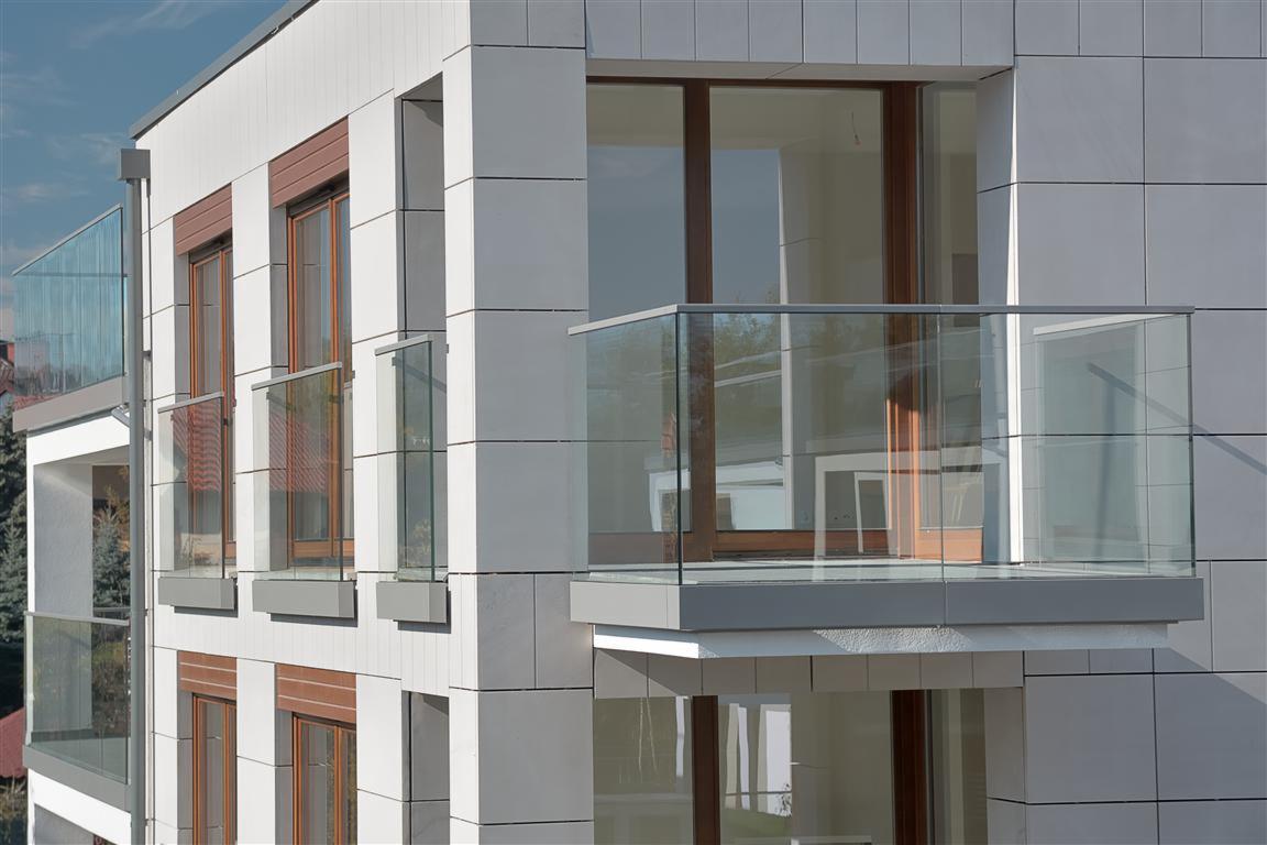 Nowoczesny budynek mieszkalny w Mosinie z balkonami z szklanymi balustradami i oknami z brązowymi ramami, wykończony płytami elewacyjnymi w odcieniach szarości i bieli.
