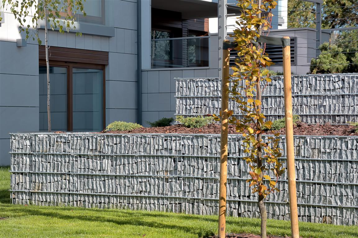 Gabionowa ściana oporowa z szarego kamienia, zabezpieczająca skarpę z roślinami, przed nowoczesnym budynkiem z szarą elewacją i balkonem.