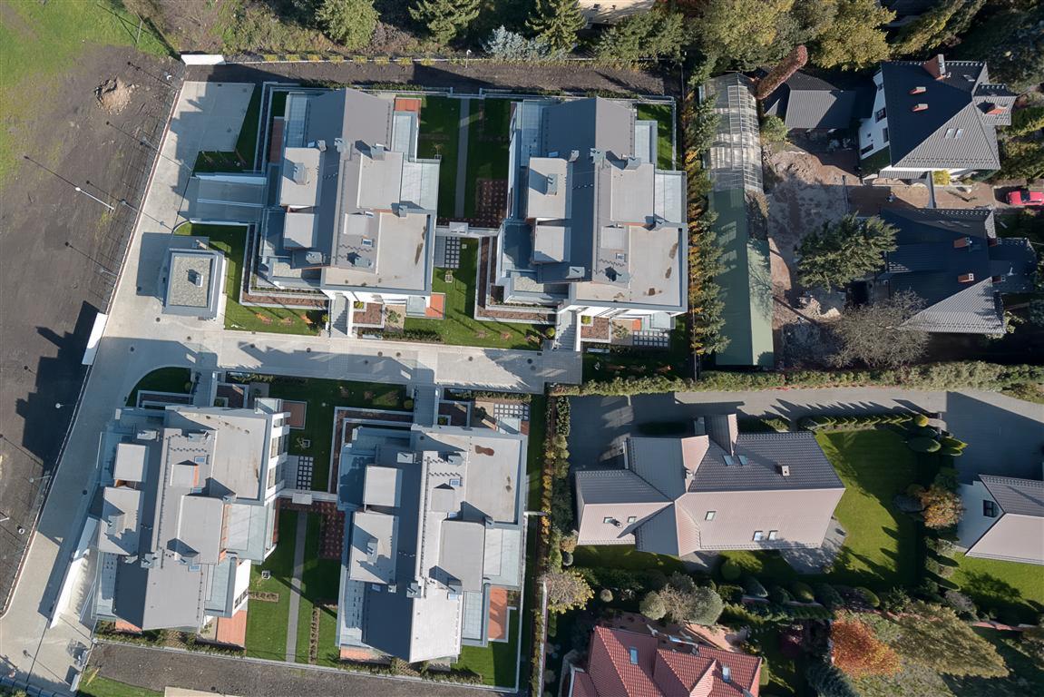 Widok z drona na osiedle nowych domów jednorodzinnych z szarymi dachami, zadbanymi trawnikami i brukowanymi ścieżkami, otoczone zielenią drzew i krzewów. Ujęcie z góry ukazuje regularny układ...