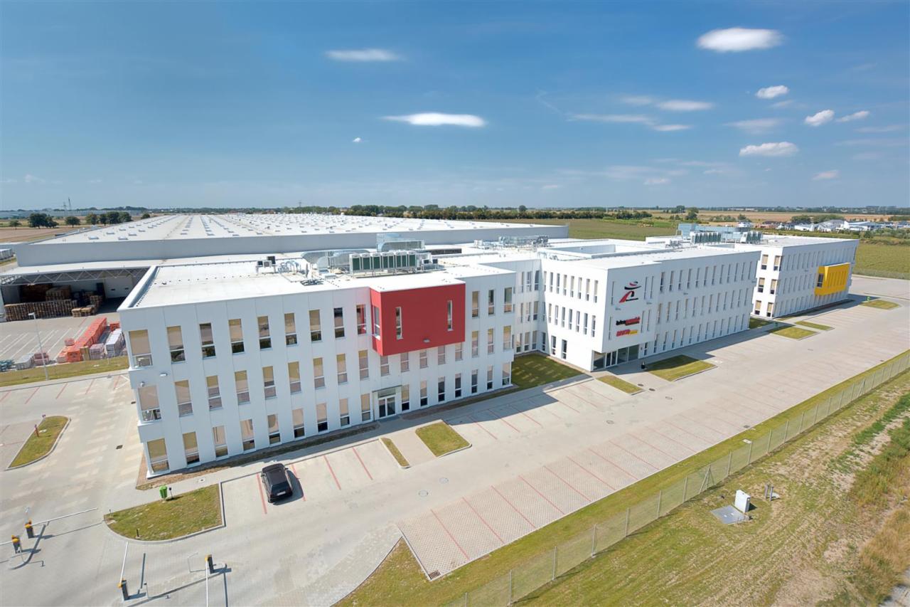 Nowoczesny budynek biurowo-magazynowy z białymi elewacjami, czerwonymi i żółtymi akcentami kolorystycznymi, widok z góry, parking, trawnik i ogrodzenie.