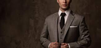 Mężczyzna w eleganckim, szarym garniturze w kratę, czarnym krawacie i białej koszuli, z białą poszetką w kieszonce, pozuje na rozmytym, ciemnym tle.