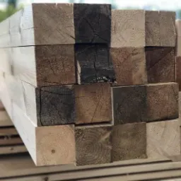 Stos kwadratowych belek drewnianych z widocznymi słojami, niekt&oacute;re belki z ciemniejszymi przebarwieniami, ułożone na palecie.