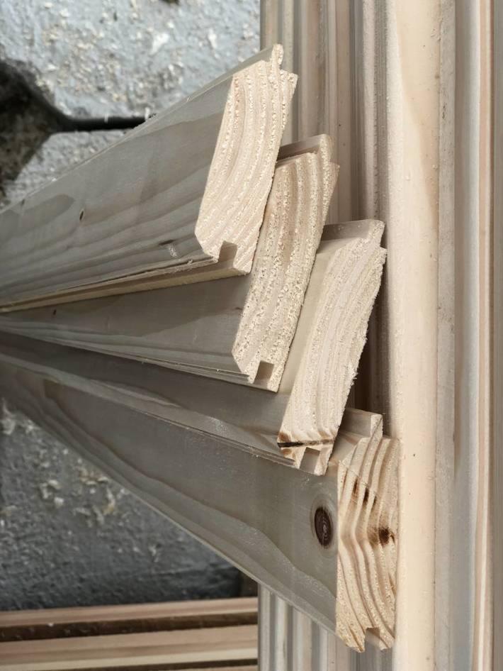 Ułożone warstwowo profile z jasnego drewna z widocznymi słojami, z charakterystycznym wycięciem na krawędzi, leżące obok pionowej, żłobionej deski.