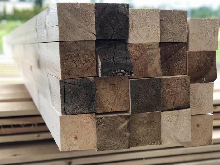 Stos kwadratowych belek drewnianych z widocznymi słojami, niektóre belki z ciemniejszymi przebarwieniami, ułożone na palecie.
