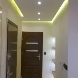 Nowoczesny korytarz z oświetleniem LED, wbudowanymi półkami z podświetleniem, dwoje drzwi w odcieniach brązu i szafa w tle.