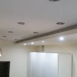 Sufit podwieszany z licznymi punktowymi oprawami oświetleniowymi, widoczny zegar na ścianie i fragmenty białych szafek.