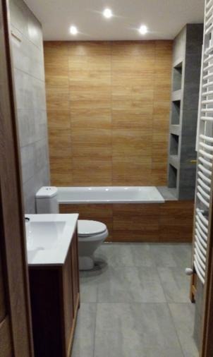 Nowoczesna łazienka z drewnopodobnymi płytkami na ścianie, białą wanną obudowaną drewnem, szarą podłogą i szafką pod umywalkę z drewnianymi frontami.