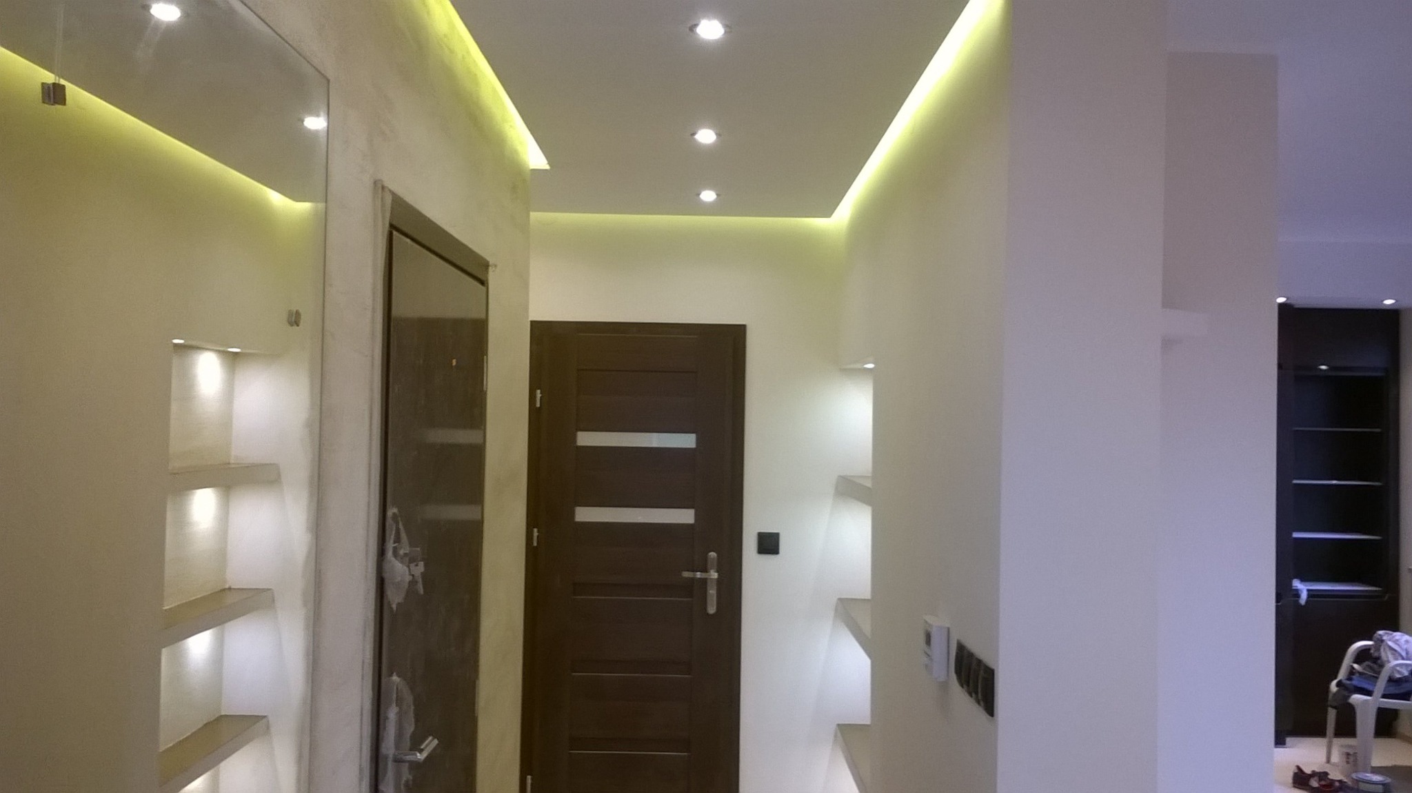 Nowoczesny korytarz z oświetleniem LED, wbudowanymi półkami z podświetleniem, dwoje drzwi w odcieniach brązu i szafa w tle.