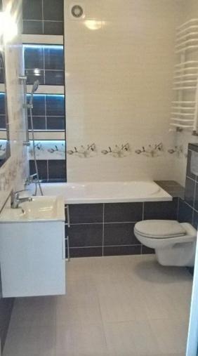 Jasna łazienka z wanną obudowaną ciemnymi płytkami, umywalką na szafce i toaletą w rogu, dekoracyjna listwa z motywem kwiatowym pod sufitem.