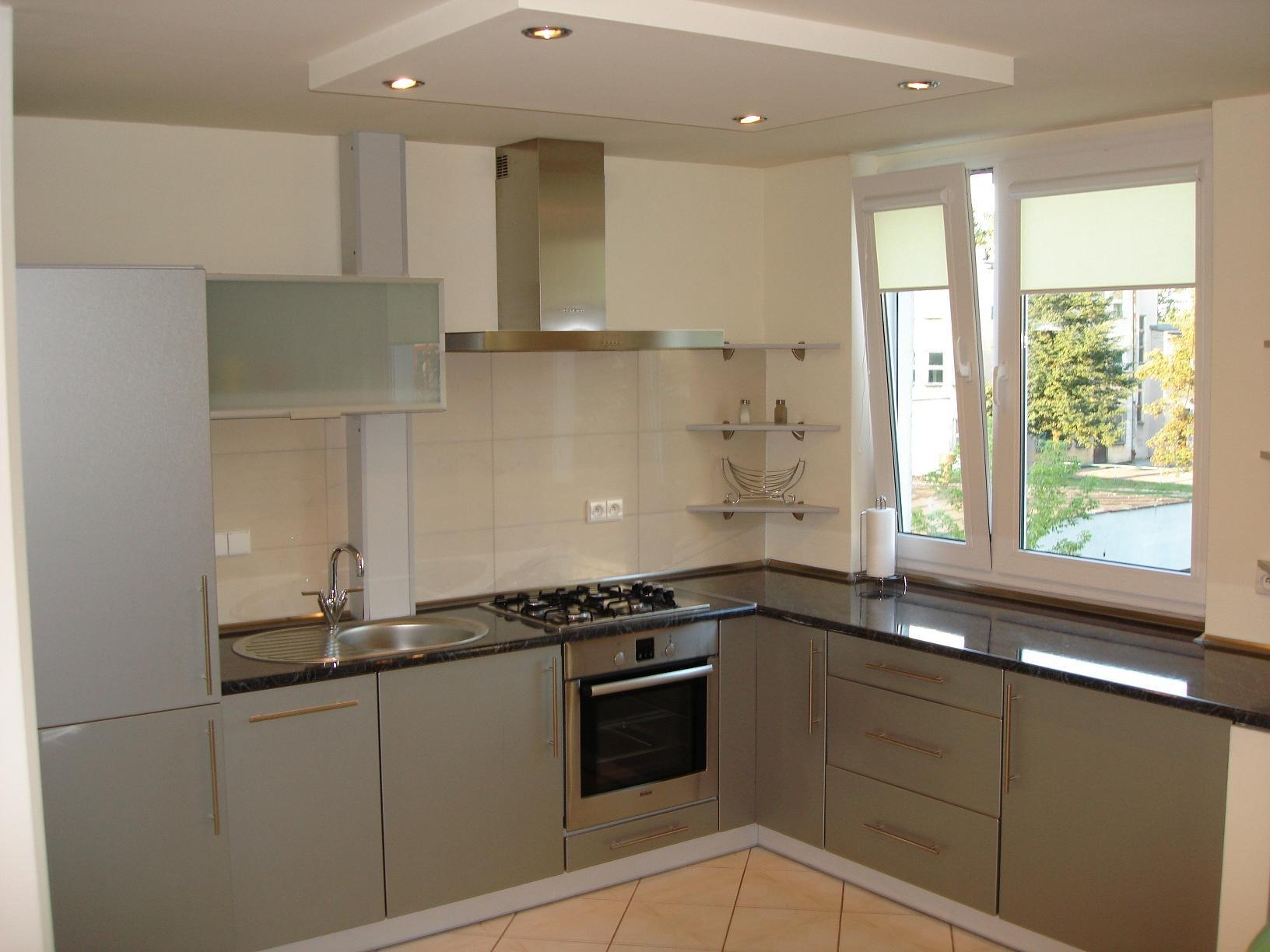 Nowoczesna kuchnia w szaro-beżowej kolorystyce z błyszczącym, ciemnym blatem, widok na okno z roletami i zabudowę kuchenną ze sprzętem AGD.