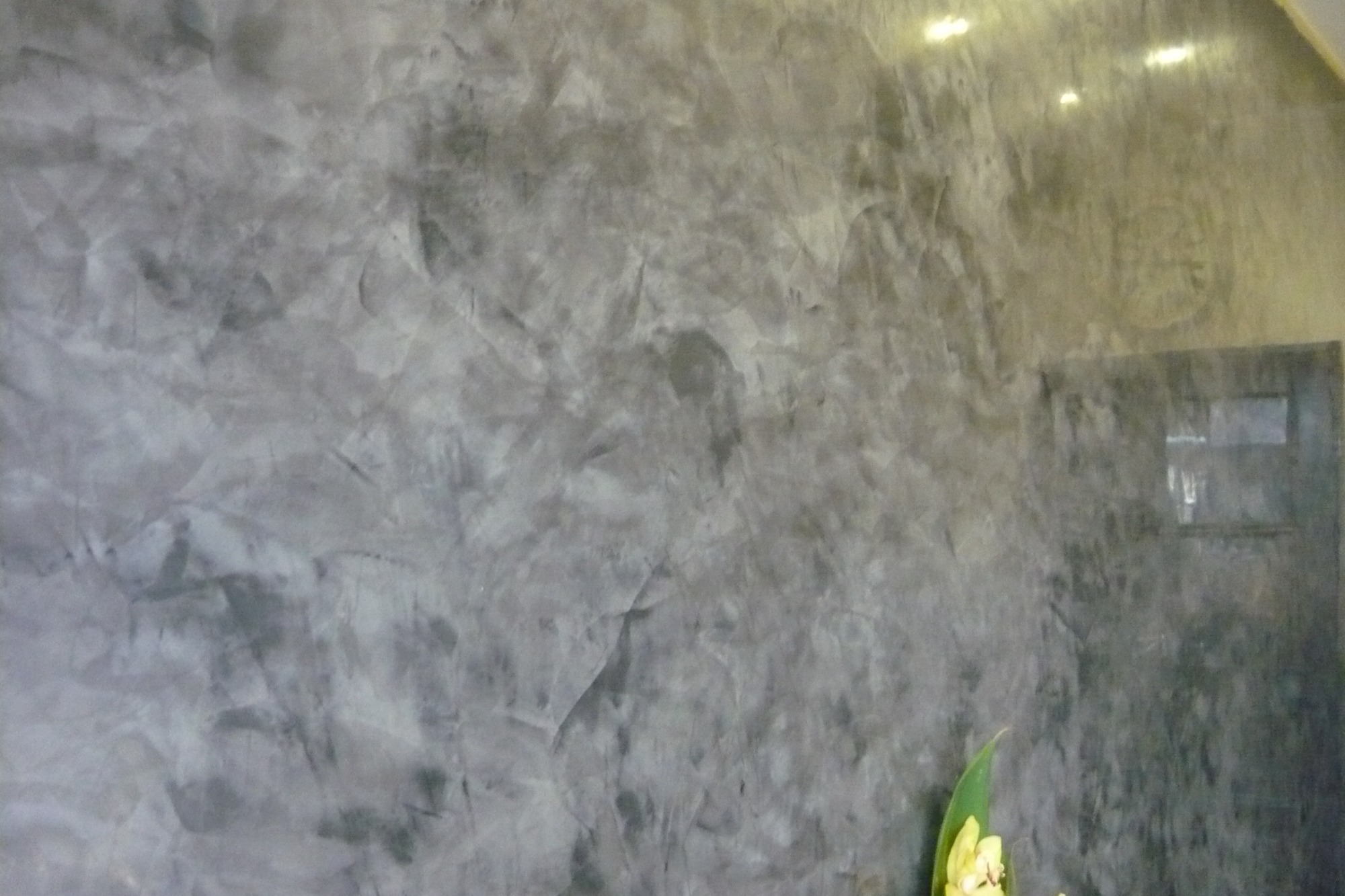 Ściana wewnętrzna z szarą, dekoracyjną fakturą imitującą kamień, oświetlona od góry, z widocznym fragmentem drzwi i rośliną w dolnym rogu.