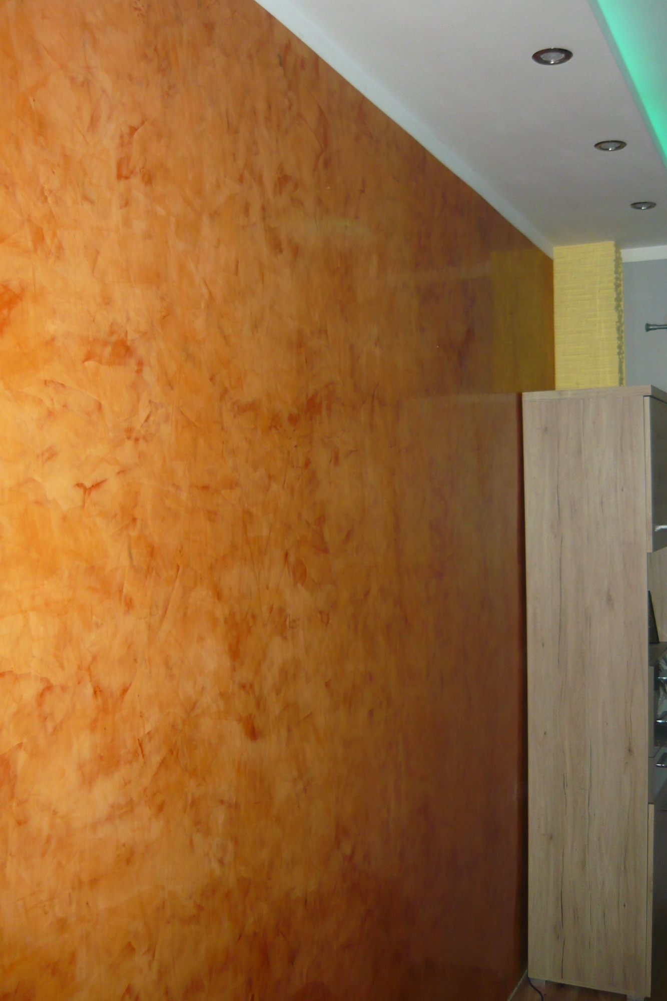 Pomarańczowa ściana z efektem dekoracyjnym, widoczne oświetlenie sufitowe i fragment mebla