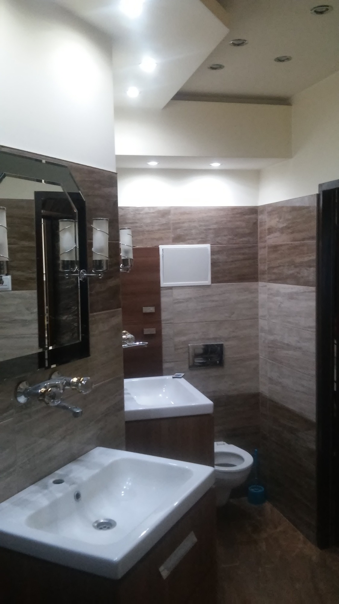 Nowoczesna łazienka z dwoma umywalkami na tle brązowych płytek imitujących drewno, podwieszana toaleta i oświetlenie punktowe.