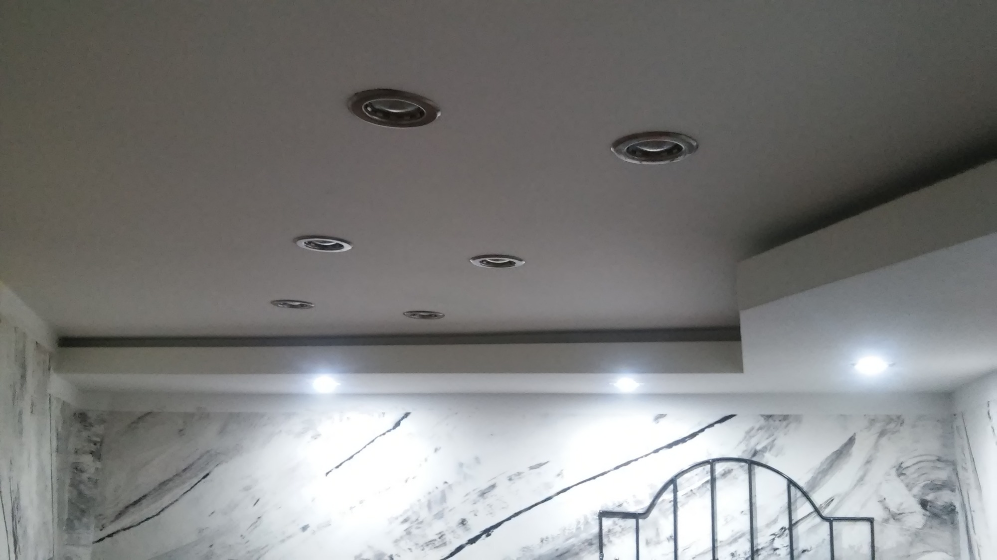 Sufit podwieszany z oświetleniem punktowym, ściany z efektem marmuru, fragment metalowego zagłówka łóżka.