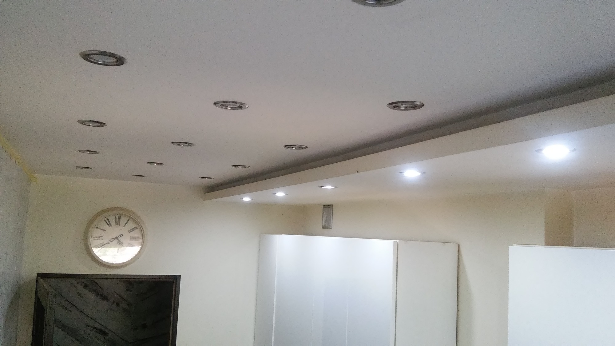 Sufit podwieszany z licznymi punktowymi oprawami oświetleniowymi, widoczny zegar na ścianie i fragmenty białych szafek.