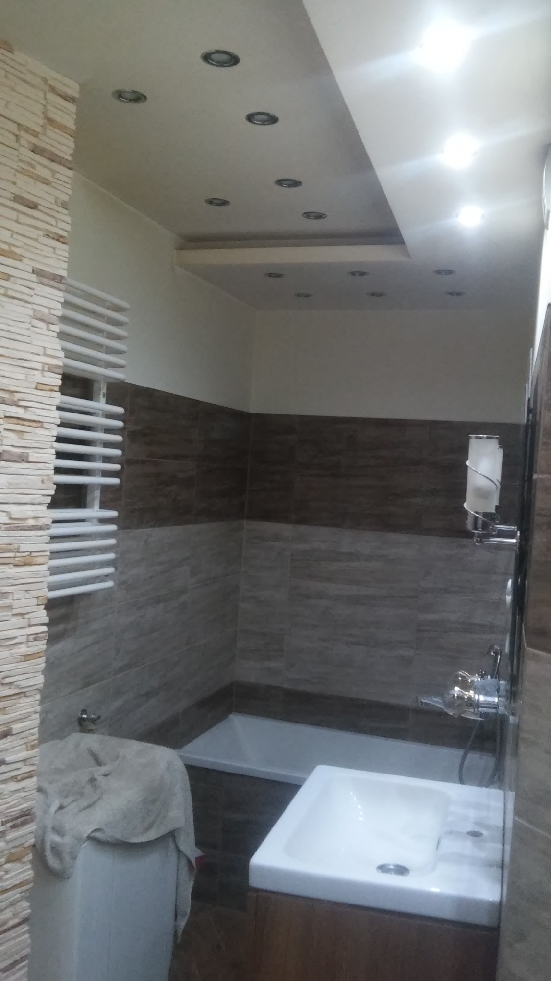 Nowoczesna łazienka z brązowo-szarymi płytkami, podwieszanym sufitem z punktowym oświetleniem, grzejnikiem drabinkowym i białą armaturą. Na pralce leży szary ręcznik.