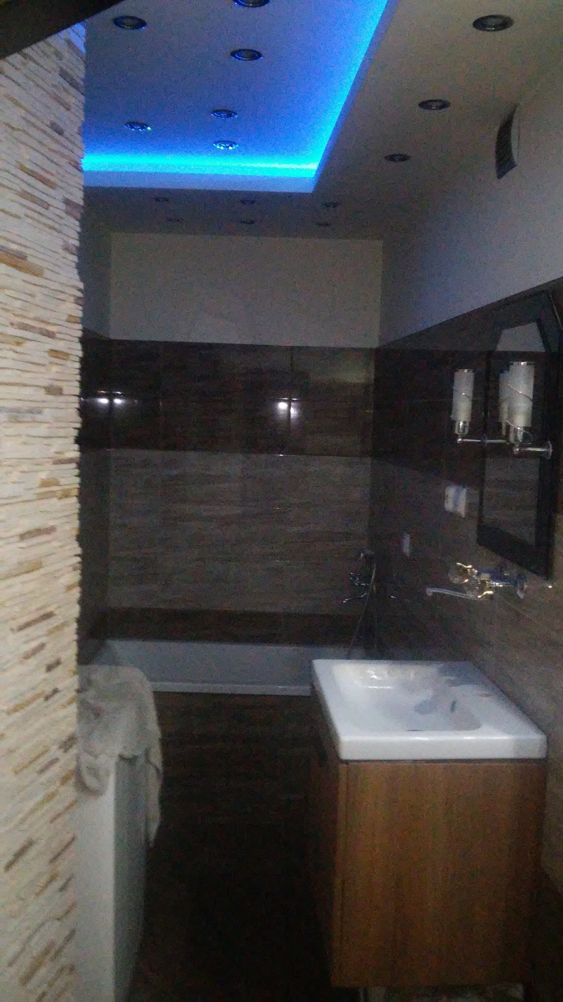 Nowoczesna łazienka z podświetlanym sufitem, ciemnymi płytkami i umywalką na drewnianej szafce, widok z perspektywy wejścia.