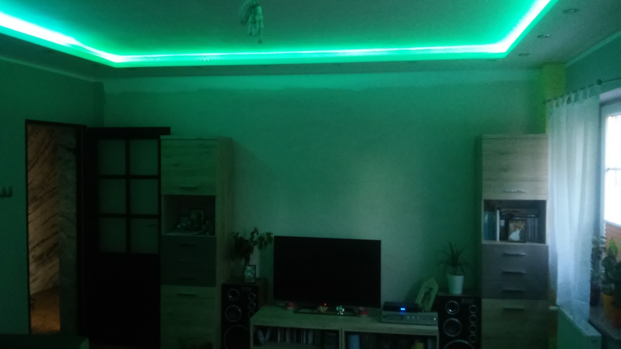 Pokój z podwieszonym sufitem i oświetleniem LED w kolorze zielonym, widoczne meble, telewizor i okno z zasłoną.