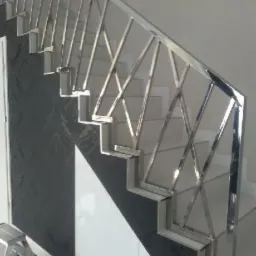 Nierdzewna balustrada schodowa o geometrycznym wzorze, widoczna z dołu, z fragmentem drabiny malarskiej w lewym dolnym rogu.