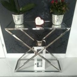 Unistal - Szklany stolik konsola z chromowanymi nogami w kształcie litery X, na blacie dwie doniczki z kwiatami i dekoracja w kształcie serca, pod blatem biała latarenka.