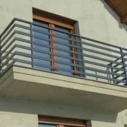 Nowoczesna, szara balustrada balkonowa z poziomych profili stalowych, zamontowana na betonowej płycie balkonu, widok z dołu na tle elewacji budynku.