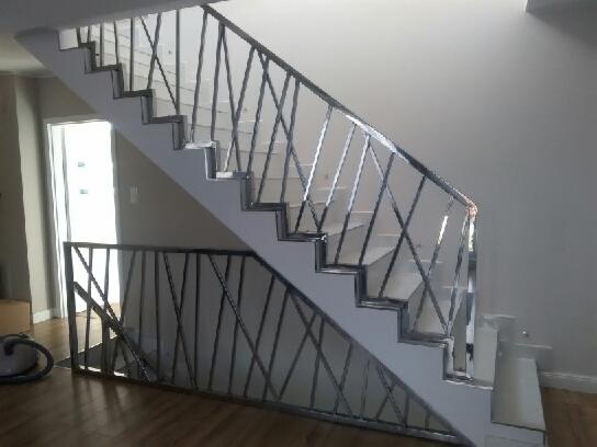 Białe schody z metalową balustradą o ukośnym wzorze krzyży, widok z dołu na podest, podłoga drewniana, jasne wnętrze.