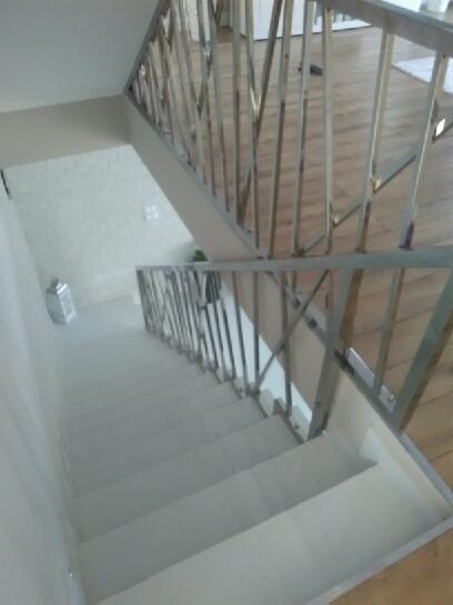 Widok z góry na schody z białymi stopniami i nowoczesną balustradą ze stali nierdzewnej z pionowymi prętami, na tle drewnianej podłogi na górnym poziomie.